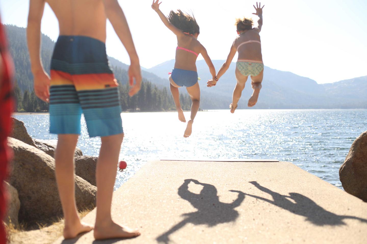 Kids jumping off a dock into a lake, sunny day, shadow visible.