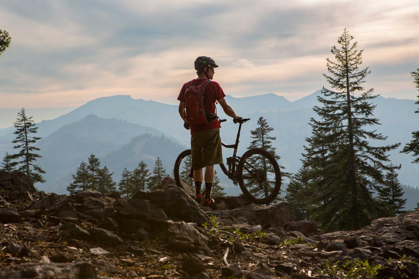 Cyclist on a rocky hilltop, overlooking mountains and pine trees at sunset.