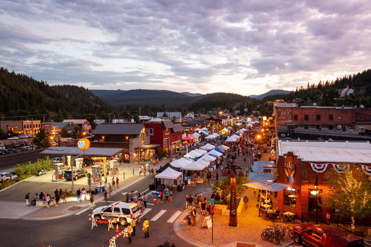 Street fair at sunset with festive lights and crowds, surrounded by mountains and shops.